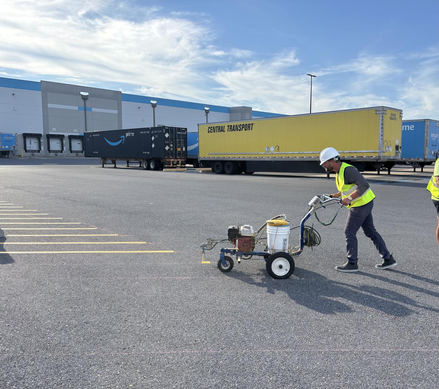 Striper in a hard hat and safety vest pushing a line striping machine past trailers at a distribution center in Carencro, LA
